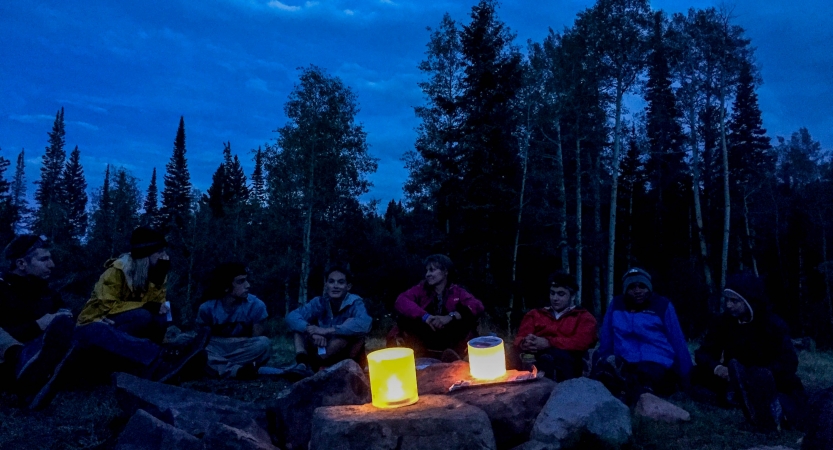 Two lanterns illuminate a group of outward bound students resting at dusk